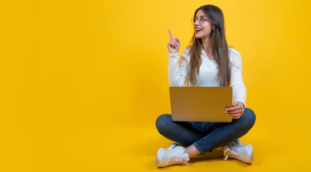 Smiling woman sitting cross-legged with laptop, raising finger in eureka moment