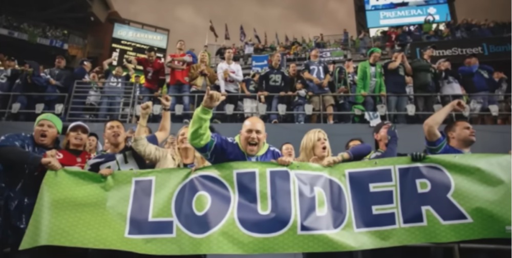 Seattle Seahawks fans holding LOUDER banner cheering in stadium stands.