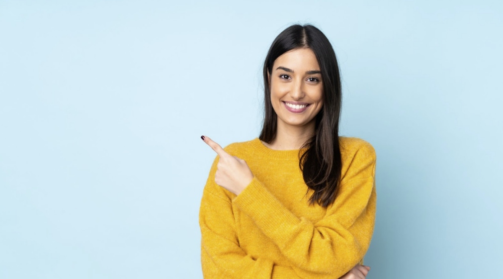 Smiling woman in yellow sweater pointing to the side on light blue background.