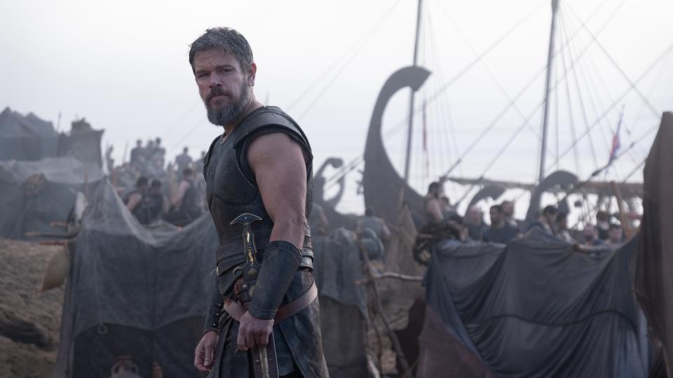 Armored warrior stands on foggy beach beside ancient ships.