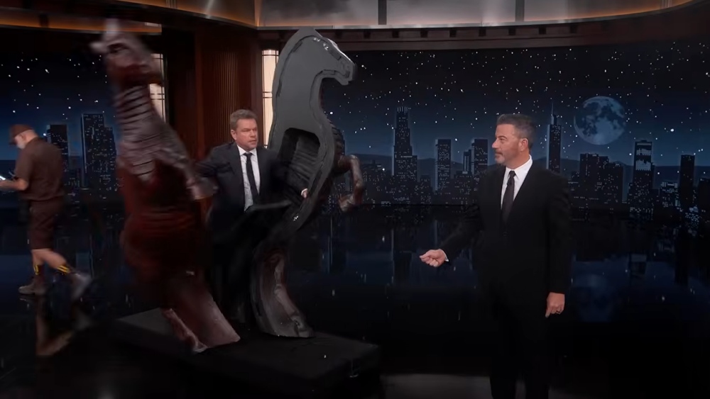 Late-night host stands beside horse sculptures on TV talk show set.