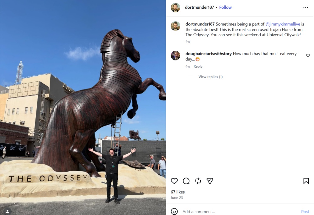 Man poses arms raised by "The Odyssey" Trojan Horse statue outdoors.