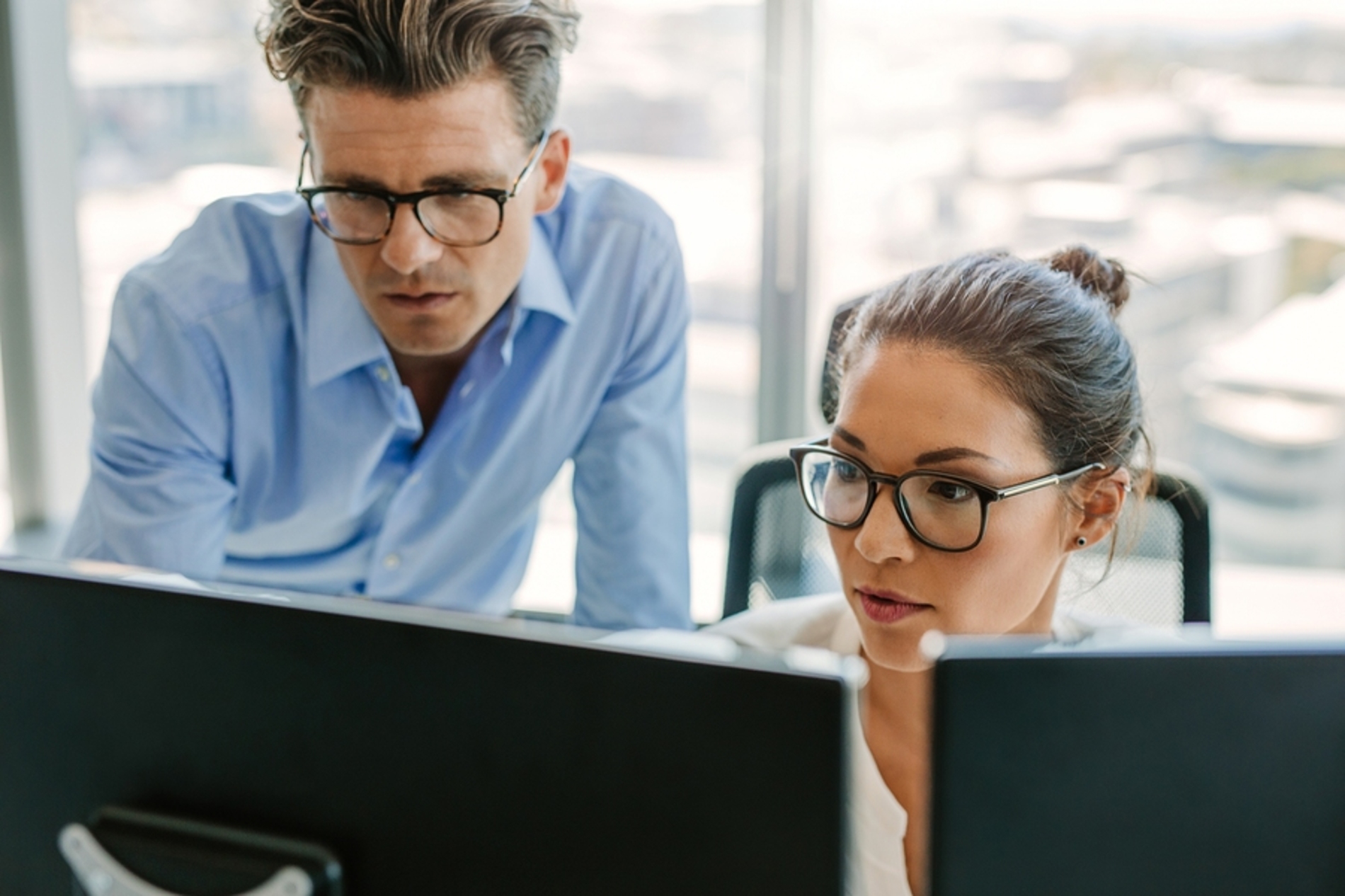 Two coworkers looking at computer monitors together. Two coworkers looking at computer monitors together.