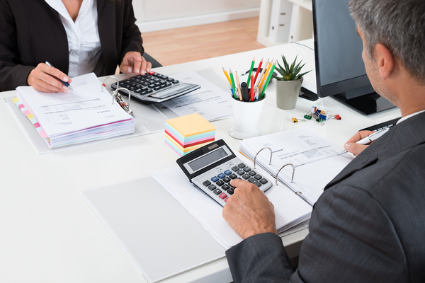 Coworkers at a shared desk with documents and calculators.