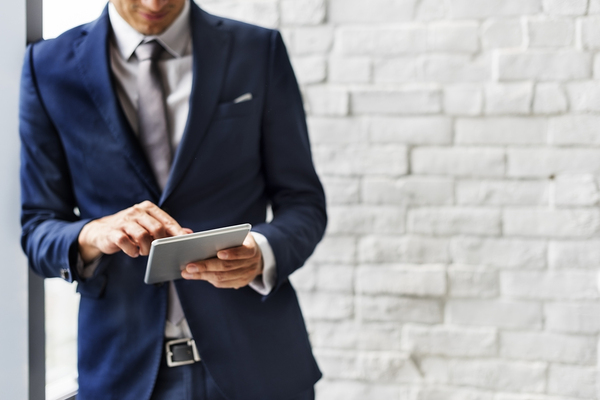 Man wearing a suit and leaning against a wall using his tablet.