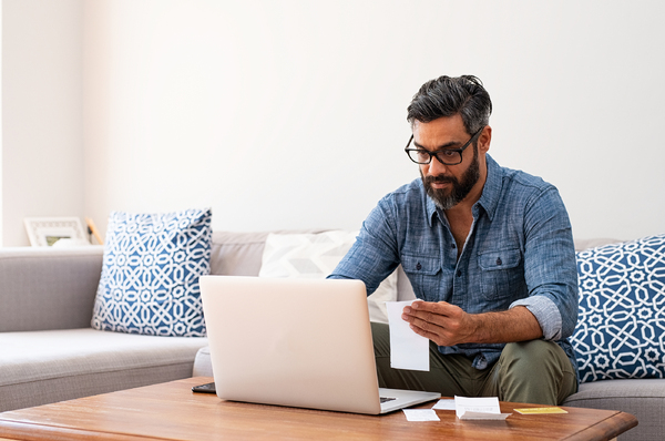 Man sitting on his couch looking at his computer screen while holding a document.