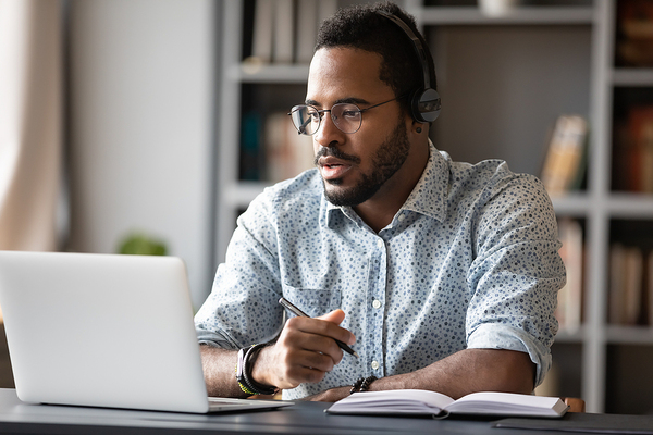 Man wearing headphones and looking at his laptop screen while taking notes.