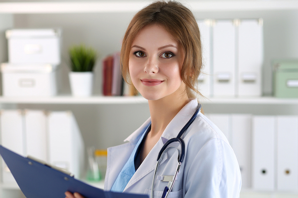 Female doctor holding a clipboard and wearing a white lab coat with stethoscope around neck smiling.