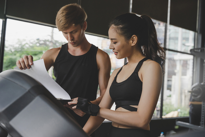 Woman working out on an exercise machine consulting a personal trainer.