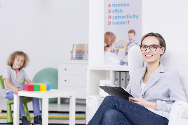 Therapist holding a clipboard with a child in the background playing with blocks.