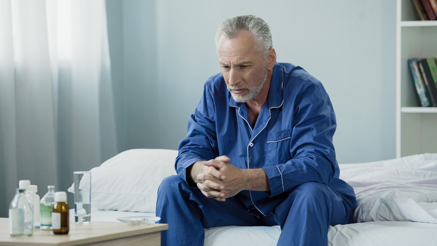 Man sitting at the side of his bed looking down.