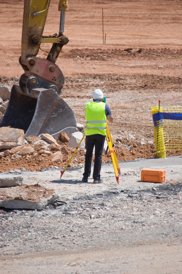 Engineer using surveying equipment at a construction site with a backhoe in the background.