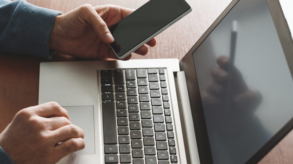 Person typing on a laptop and referencing their mobile phone.