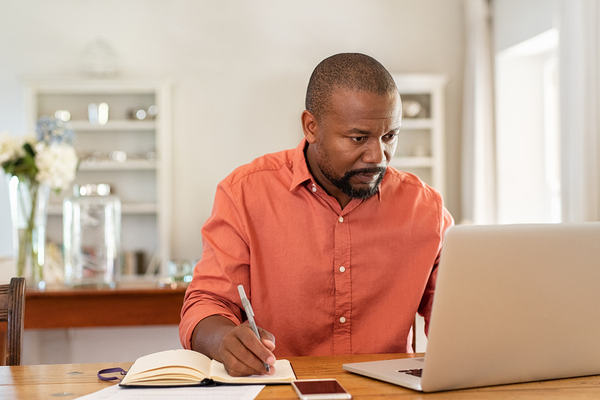 Person looking at their laptop screen while writing in a notebook.