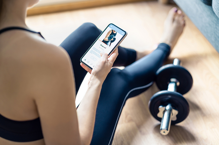 Woman sitting on the floor look at at phone with dumbbells next to her.