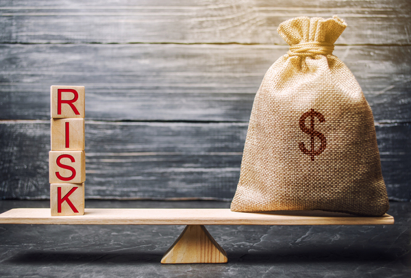 Wooden scale leveled by blocks spelling risk, and a burlap bag labeled with a dollar sign.