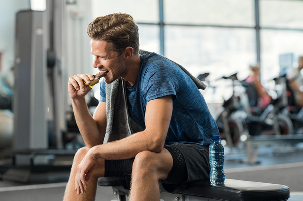 Man sitting on a bench at a gym eating an energy bar.