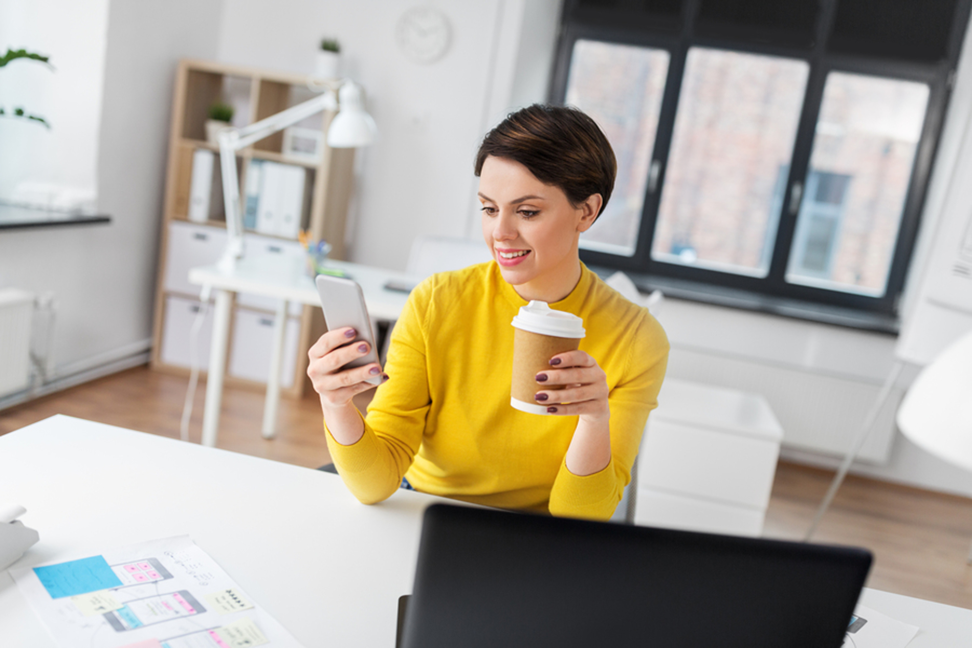 Young woman sitting at her desk looking at her phone and drinking a cup of coffee.