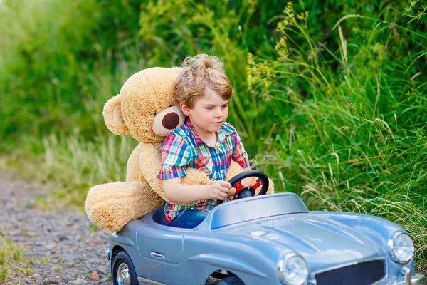 Young child driving a kid car with a teddybear behind him.